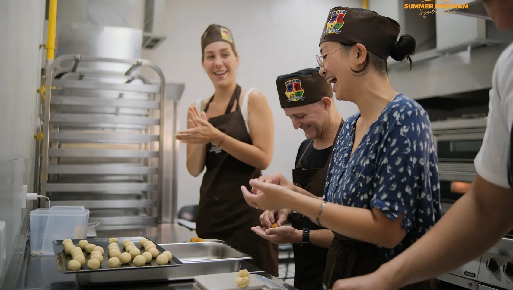 Three students laughing while shaping dough beside a full baking tray