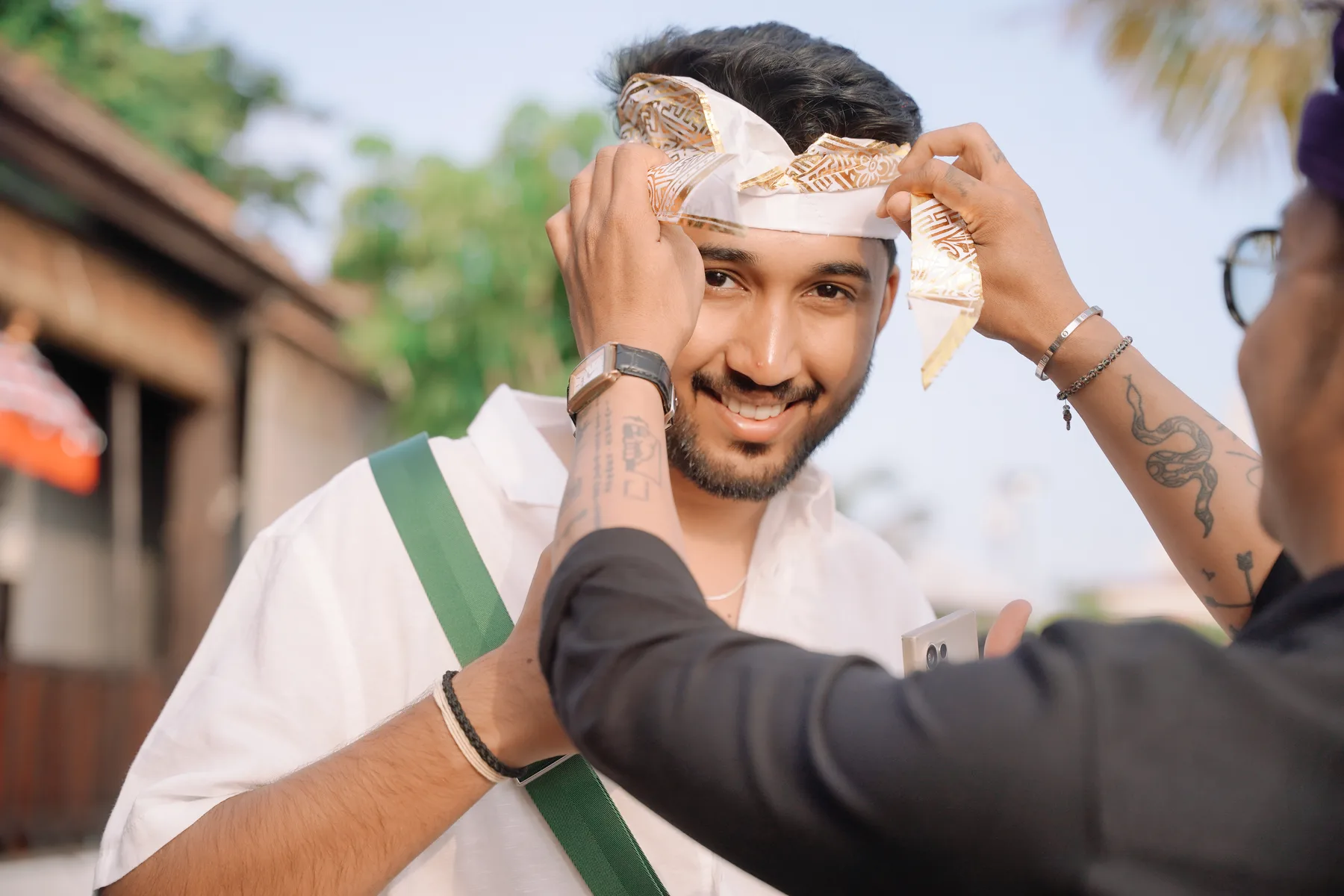 A student smiling as another person ties a gold-patterned headscarf for him