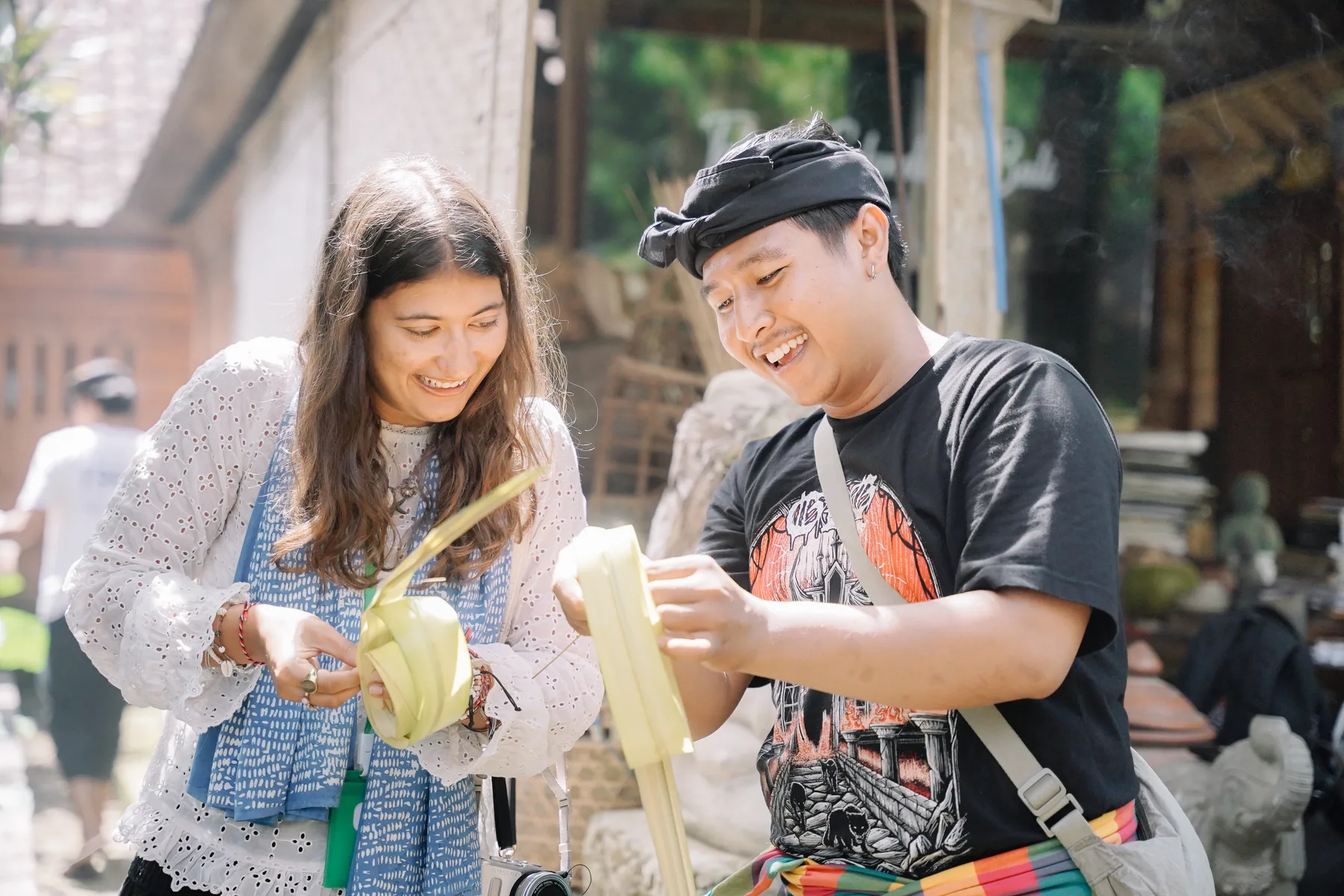 A student and a Balinese guide folding palm-leaf offerings together at Omah Blumbungan, both smiling