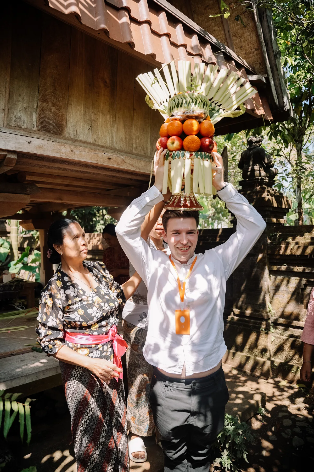 A student holding an elaborate fruit offering overhead at Omah Blumbungan, helped by a Balinese woman