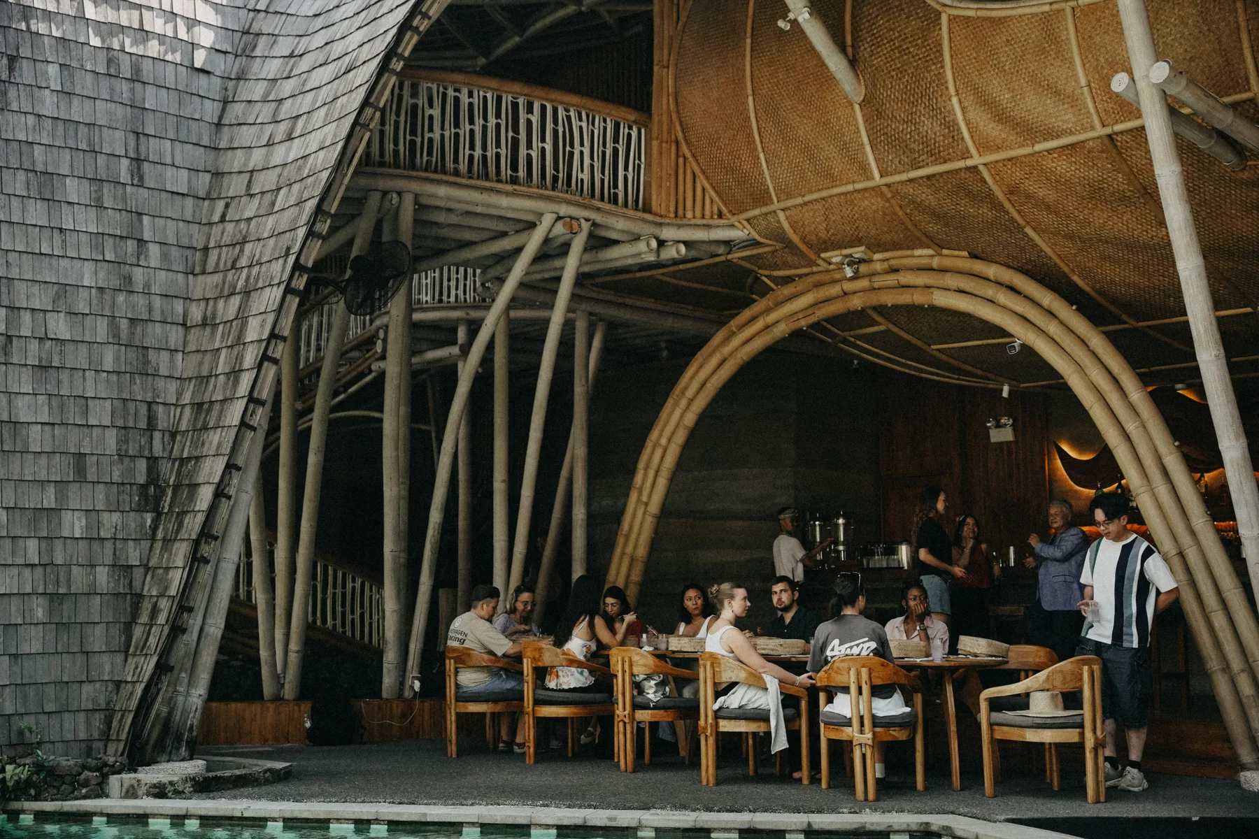 Guests seated at wooden tables inside a bamboo-framed restaurant with arched beams and a shingled roof at Nuanu