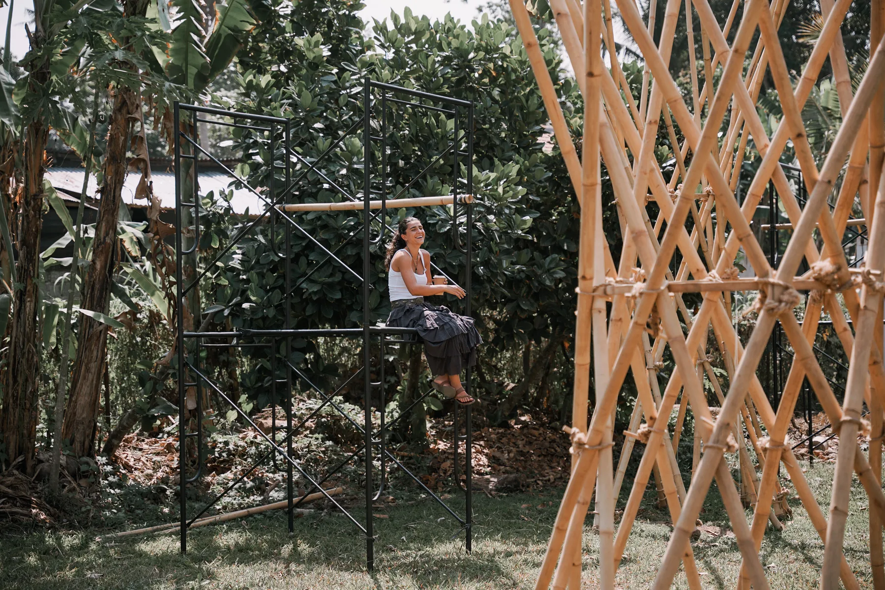 A student sitting and laughing on a steel frame beside a bamboo cross-braced structure in a garden