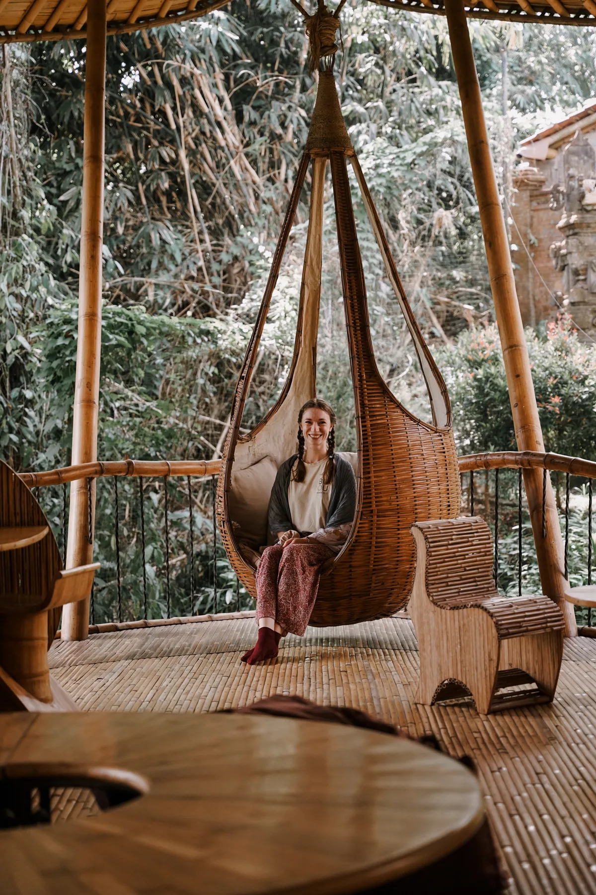 A student smiling in a large hanging woven bamboo swing chair under a thatched shelter at BambooU