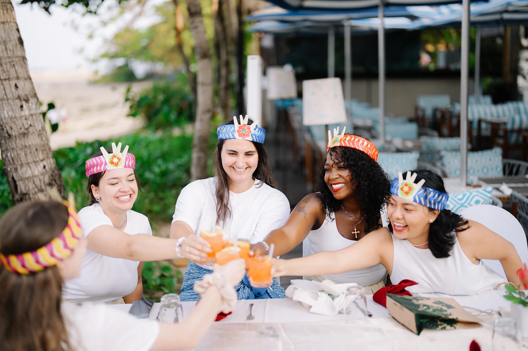 Students in white outfits and colorful woven crowns laughing while clinking drinks together