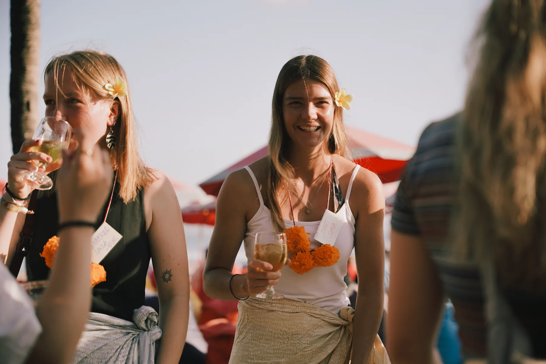 Two students toasting with drinks on the beach, one wearing a name tag that reads Sport Joana