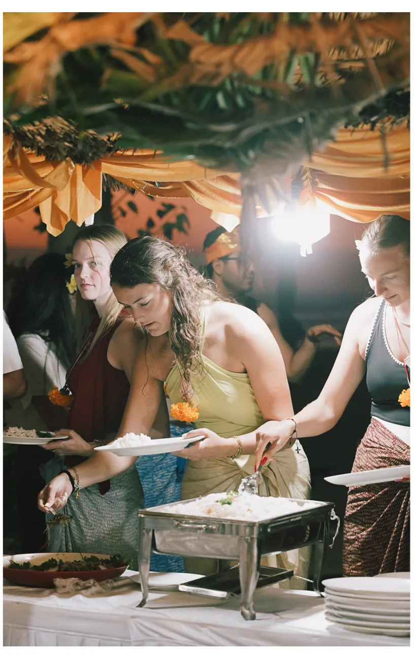 Students serving themselves from a buffet under a palm-leaf roof